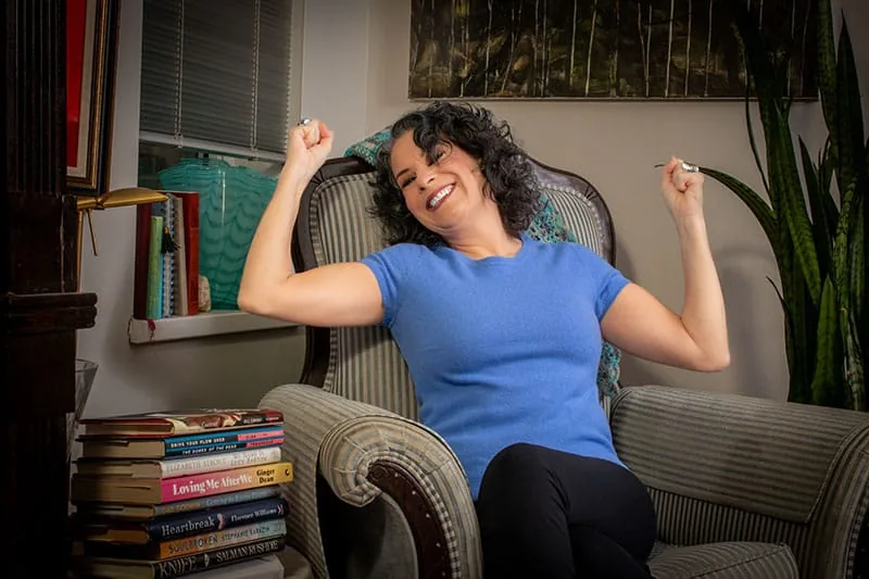 Jodee Blanco smiling and stretching in an armchair beside a shelf of books